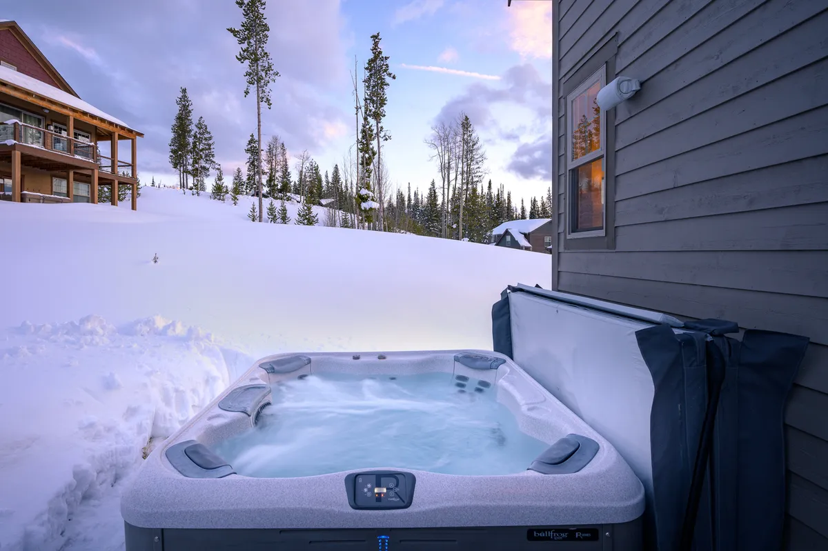 A serene exterior winter scene featuring a bubbling hot tub on a wooden deck, surrounded by snow-covered ground and tall trees, with the house visible nearby. Steam rises from the hot tub, suggesting warmth and relaxation after outdoor adventures.