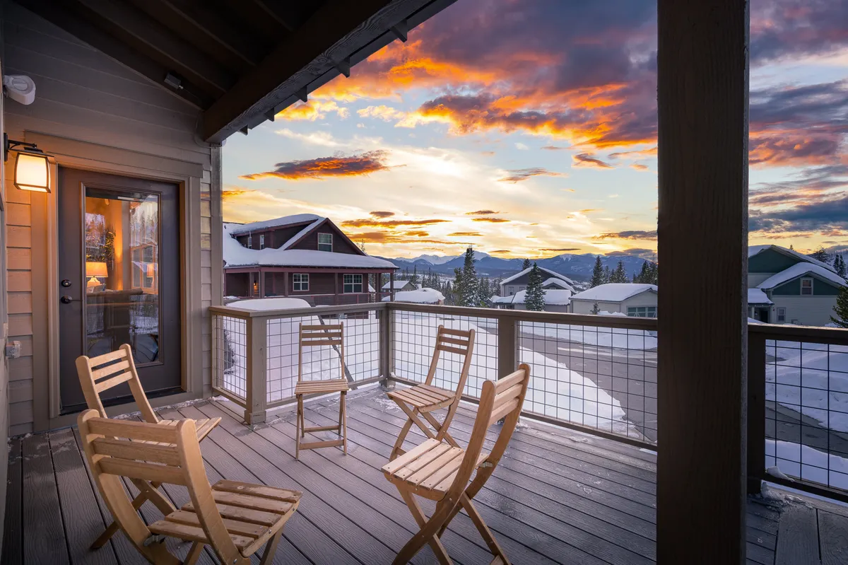 An inviting deck with wooden chairs overlooks stunning mountain views beneath a dramatic sunset sky. The peaceful setting is perfect for relaxing with friends or enjoying a morning coffee while surrounded by natural beauty.