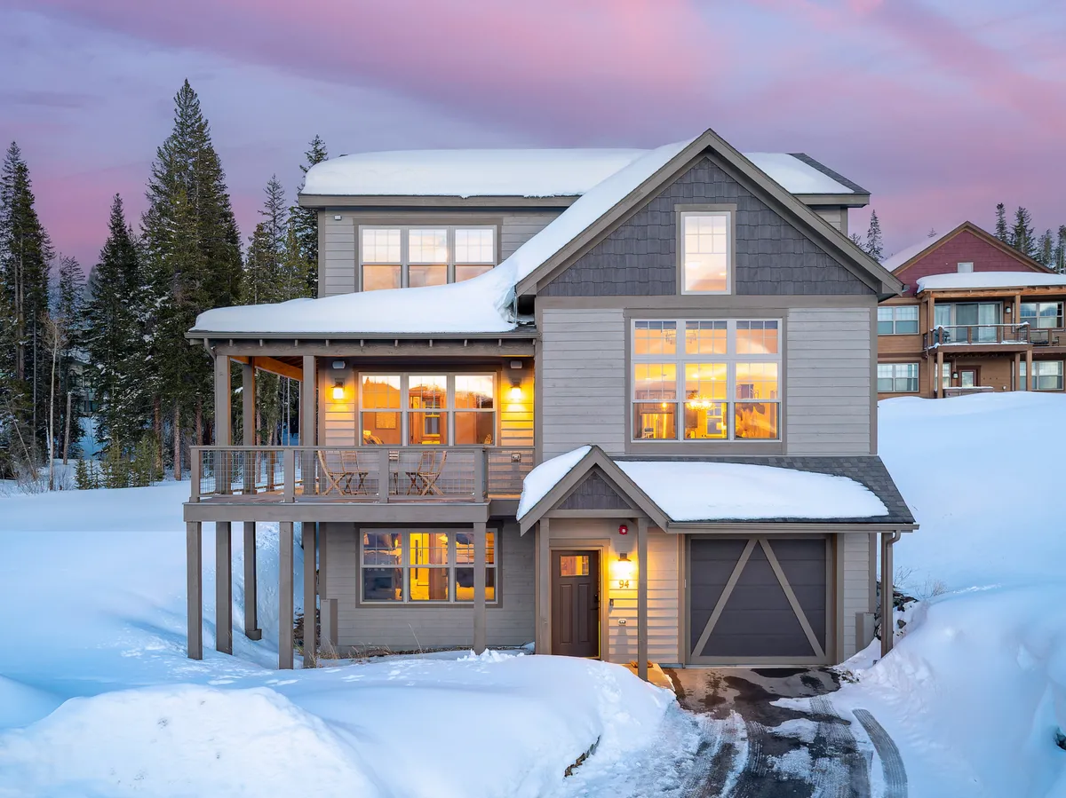 The image showcases the chalet's striking mountain architecture with expansive windows aglow from interior lights, a snow-blanketed porch suggesting cozy winter evenings, and serene mountain surroundings. The visual invites thoughts of warmth and togetherness, set against the peaceful beauty of Fraser, Colorado.