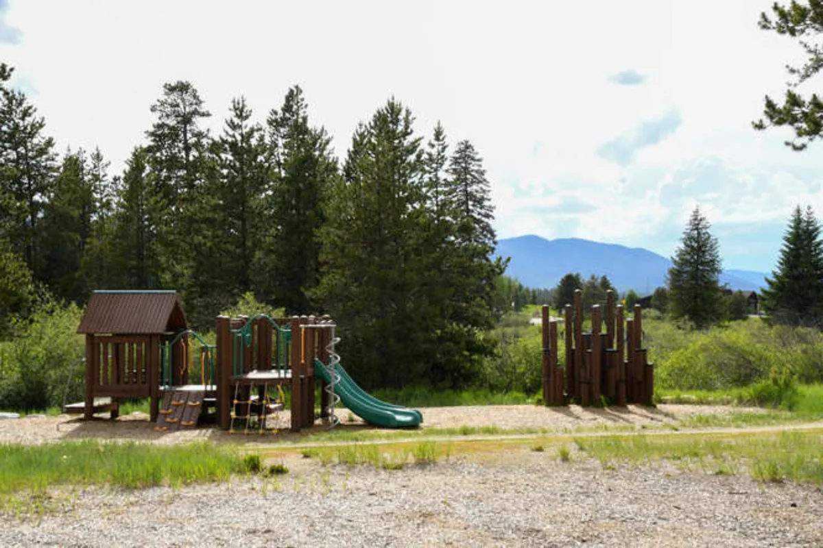 A vibrant outdoor playground surrounded by trees and set against a stunning mountain backdrop. Children play on various playground equipment, while families gather for picnics and nature walks in the joyful, adventurous atmosphere.