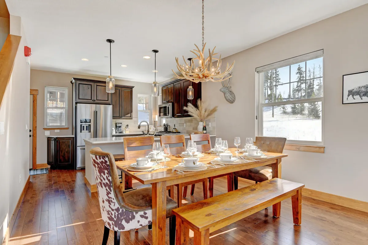 A beautifully set dining table beneath an antler chandelier, seamlessly connected to a modern kitchen with stainless appliances. Large windows frame scenic views, enhancing the inviting, social atmosphere ideal for both casual and celebratory gatherings.