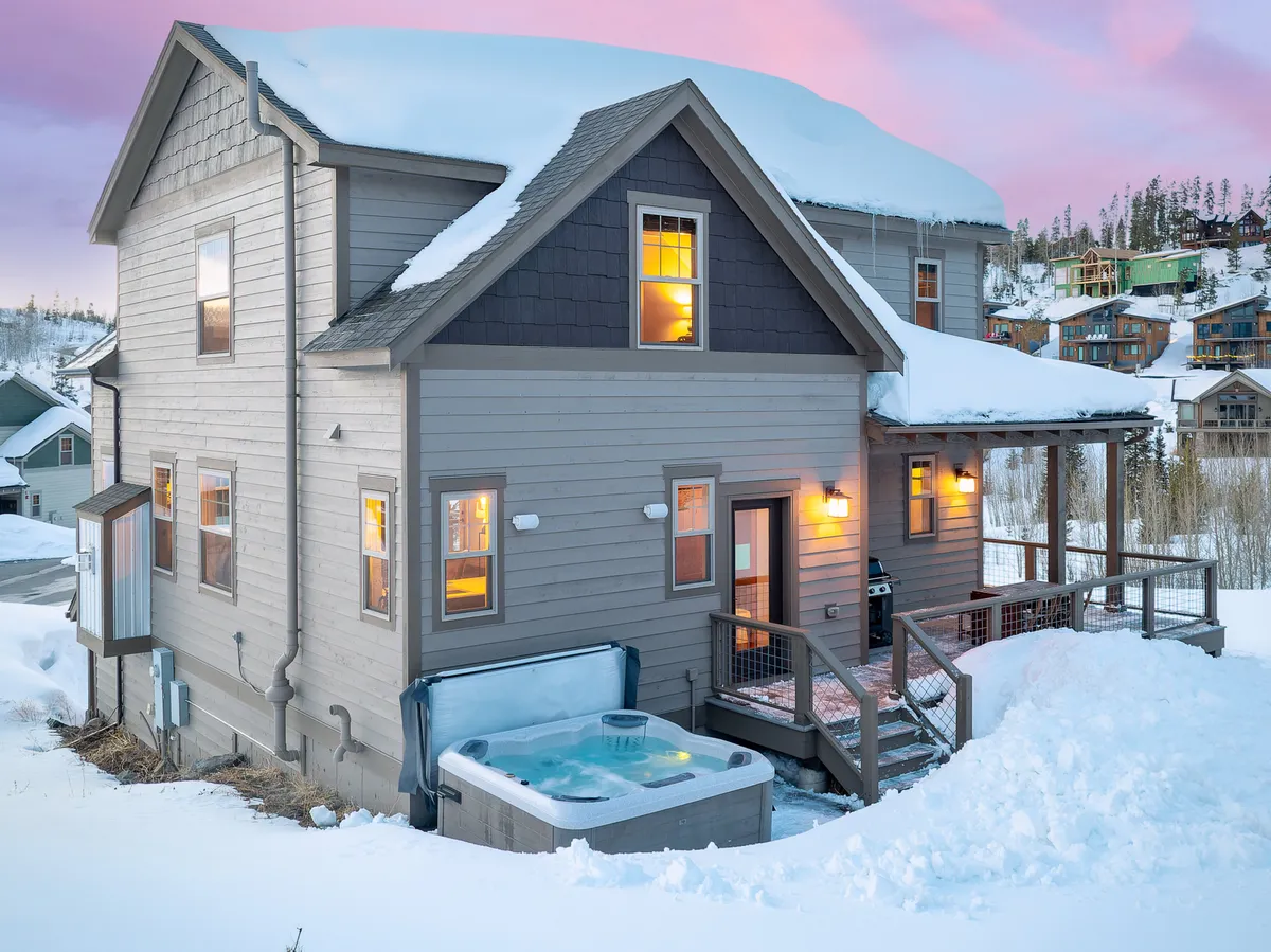 An inviting exterior shot of a private hot tub on a wooden deck, with snow covering the ground and windows glowing warmly in the background. The scene conveys relaxation after a day of winter adventure, with opportunities for stargazing and enjoying mountain views.