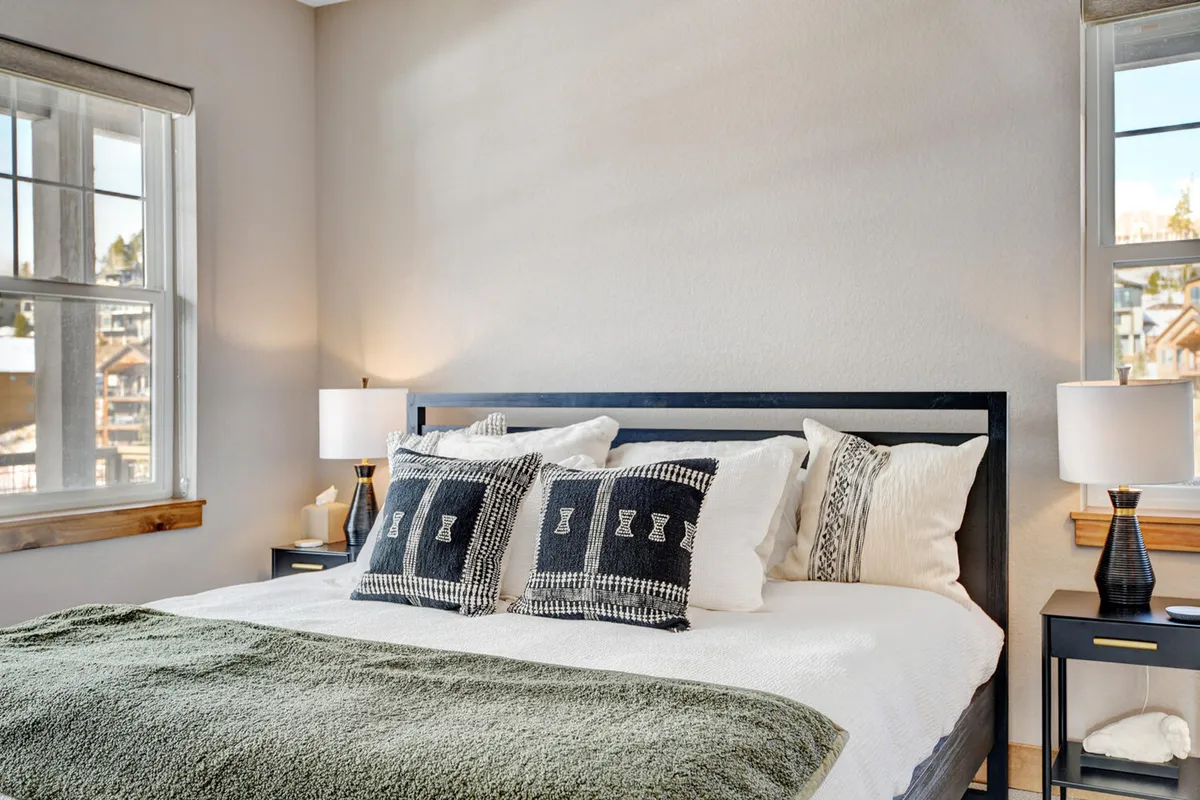 This image showcases a serene bedroom with a bed adorned with decorative pillows, nightstands with lamps on both sides, and large windows that flood the room with natural light. The setting exudes comfort and tranquility, making it an ideal private retreat after an adventurous day.