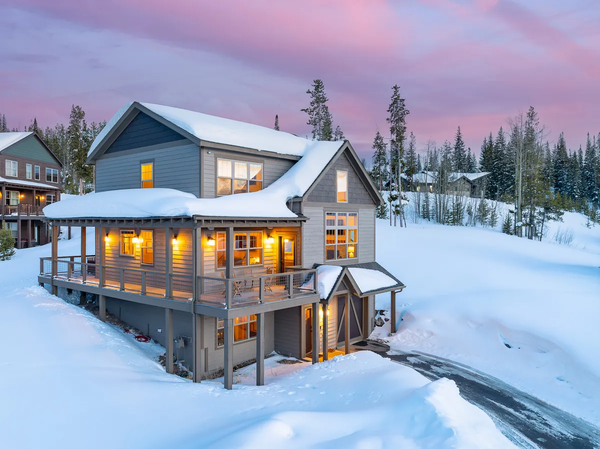 A snow-covered chalet exterior glows with warm light streaming from large windows. The spacious deck area is visible, inviting guests to relax and enjoy the mountain setting. The surrounding landscape is serene, with soft snow and distant peaks hinting at adventure and privacy.