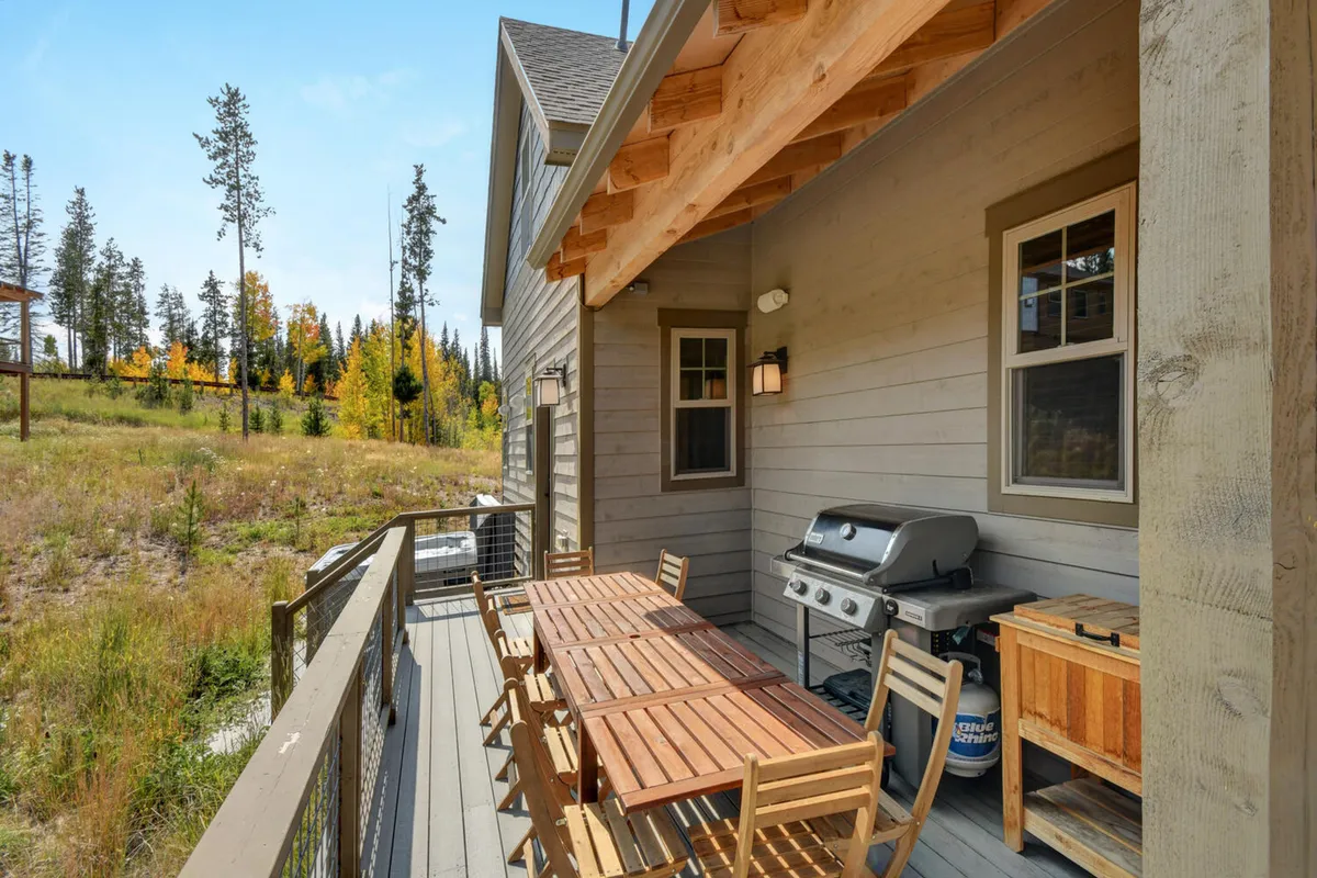 An exterior shot of a wooden deck featuring an outdoor dining table and a gas grill, framed by surrounding trees and open sky. The relaxed, nature-connected setting is perfect for alfresco dining and barbecues with mountain vistas.